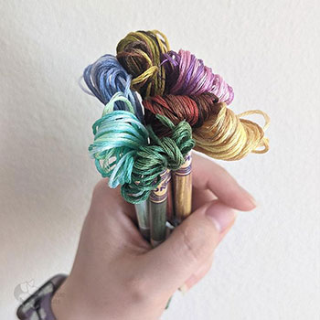 A hand holding 7 skeins of DMC flosses
