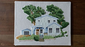 A large cross-stitch piece of a Texan home with tall trees in the background, grass in the foreground and a carport to the side.