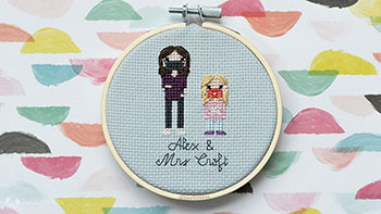 A 4in embroidery hoop with an adult (teacher) and child (student) completed in cross-stitch, both wearing teeny little face masks