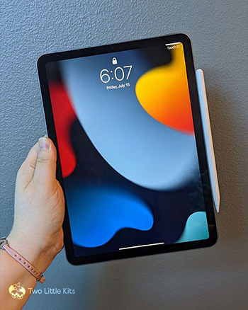 Kate's hand holding up an iPad Air (Gen 5) against a blue-grey wall. There is an Apple Pencil attached to the side and the screen is on showing a colourful background