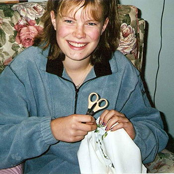 A scanned photograph of a young, teenage Kate doing hand embroidery. She’s sitting in an old-looking floral chair wearing a fuzzy, warm jumper, smiling. She has in one hand a piece of cloth that you can slightly make out embroidery work on it and a pair of scissors in the other hand.