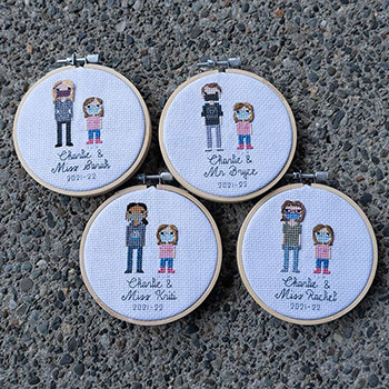Four 4in embroidery hoops of crosss-stitched pieces that all include one adult and the same little girl across them all. They are stitch people portraits and all say 'Charlie &' and then her teacher's name with the date '2021-22' underneath. They are all laid flat on a stony floor.