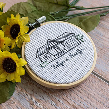 A small backstitched piece of a house outline with bushes that reads 'Robyn & Jennifer' underneath it.