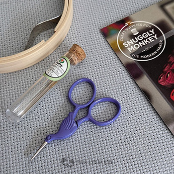 An array of stitching supplies laid out: A tension-based embroidery hoop, a vial of Tulip branded needles, small purple stalk snips and some grey-blue Aida fabric.