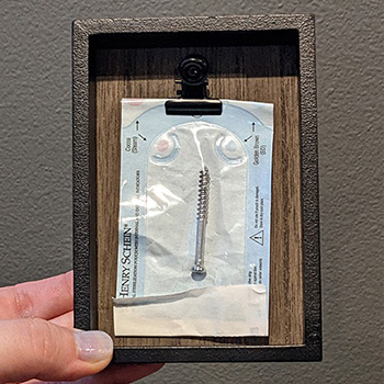 A picture of a surgical screw in a sterile packet that is clipped into a little shadowbox frame.