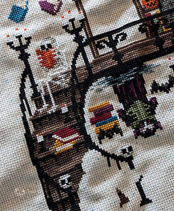 An angled photo of a section of a large cross-stitch. You can see a skeleton reading an orange book and Dracula reading a green book. They are amongst a spooky staircase.