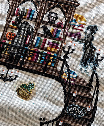 An angled photo of a section of a large cross-stitch. There is a ghost librarian with a large stack of books, a raven, a dismembered head and a headless body holding its head in its hand.