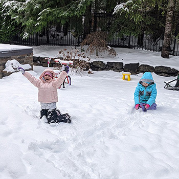 Two kids in a backyard that's covered in thick snow wearing winter clothing. They are both having a lot of fun.