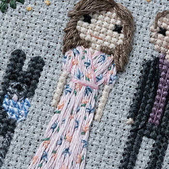 A close-up photograph of a cross-stitch & hand embroidered woman wearing a pale pink, floral dress. She has brown hair and is next to a (hinted) stitched dog and man.
