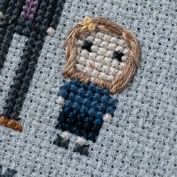 A cross-stitch and hand embroidery depiction of a little, blonde girl. She is wearing a blue and black dress, teensy little grey shoes and a yellow bow in her hair.