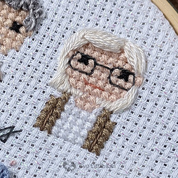 A close-up photo of a cross-stitched woman with white/blonde hair. She has dark framed glasses on and wearing a white top and brown cardigan. Her hair is pinned to the side.