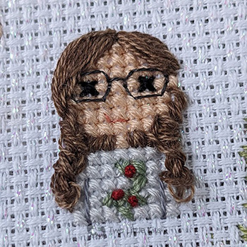 A close-up photo of a cross-stitched girl with beautiful hand-embroidered, wavy brown hair. She is wearing a grey top with mistletoe on it and has large, dark-framed glasses on.