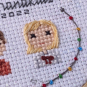A close-up photo of a cross-stitched little blonde-haired girl. She is wearing a top that has a red bow tied on the front and you can also see a string of hand embroidered 