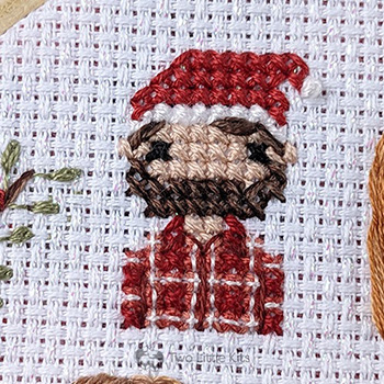 A close-up photo of a cross-stitched man wearing a santa hat. He has brown hair and facial hair. He is also wearing a buffalo-plaid shirt which has been meticulously hand stitched.