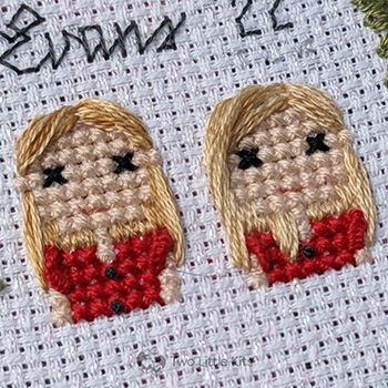 A close-up photo of two cross-stitched girls, both with shoulder-length blonde hair. They are wearing matching red dresses with frilly shoulders and black buttons down the front.