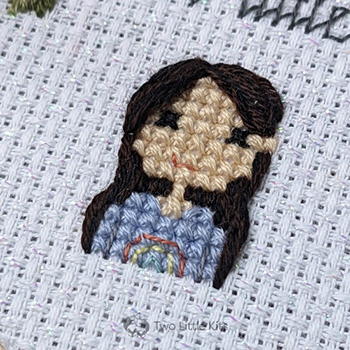 A close-up photo of a cross-stitched girl with long, almost-black hair. She is wearing a purple long-sleeved top that has a pastel rainbow on the front.