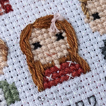 A close-up photo of a cross-stitched little girl who has bright auburn hair. She is wearing a red top that has teensy crosses on it and a cute pink bow in her hair.