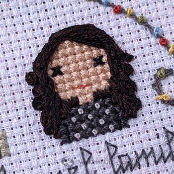 A close-up photo of a cross-stitched woman with lots of almost-black, very curly hair, parted deeply on the side. She is wearing a black top which has a dotted pattern on the front.