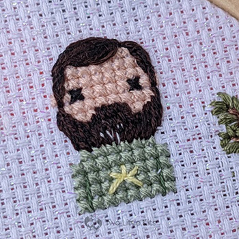 A close-up photo of a cross-stitched man. He has lots of dark brown facial hair that, at the chin -only, is speckled with salt & pepper colouring. He is wearing a light green top with a yellow star on the front.