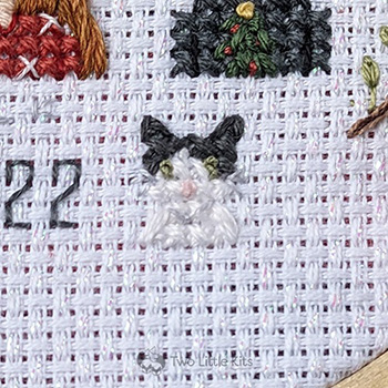 A close-up photo of a tiny little cross-stitched cat. The colouring is mostly-white with dark-grey ears and temples. It has a cute pink nose, green eyes and faint white whiskers.