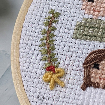 A close-up photo of hand-embroidered branch of pine needles, red berries and a yellow bow at the bottom.