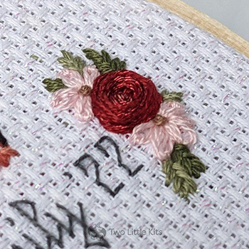 A close-up photo of hand-embroidered, small floral arrangement. It includes a large red flower (which has sparkley floss mixed in with it), pink flowers and some teensy green leaves.
