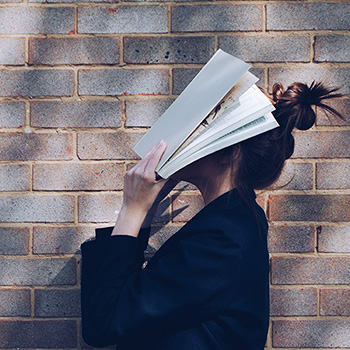 Photograph of an unknown woman standing infront of a large brick wall. She is wearing a dark blazer and is holding an open book on top of her face. Her messy bun is poking out from the top.