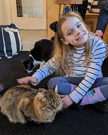 A photograph of a blonde-haired girl with cat a ear headband and drawn-on cat nose and whiskers is sitting next to a long-haired cat. She is pulling a silly smile while she pets the cat.