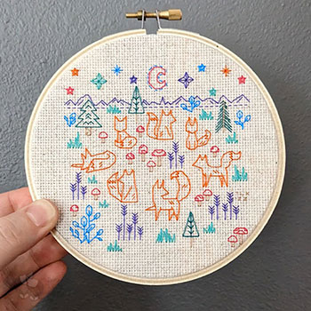 A photograph of a 5in embroidery hoop with a completed blackwork piece. It shows an imaginary scene with 7 foxes surrounding some mushrooms. Around them are more mushrooms, trees, grass, foliage and in the 'background' is mountains and the moon.