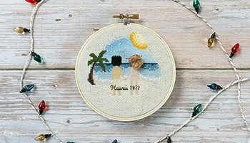 A flat-lay photograph of a cross-stitch couple portrait in a 4in embroidery hoop. The stitch depicts a man and woman standing on a beach (with palmtree and sun in the design) however, they are both naked. You can just make out underwear tan lines and butt-cracks. The woman's hair is stitched up to look like a bun. Around the hoop is a string of fake christmas lights.