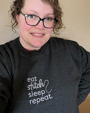 A selfie of Kate wearing a charcoal-coloured jumper that says 'eat, stitch, sleep, repeat' embroidered on the front.
