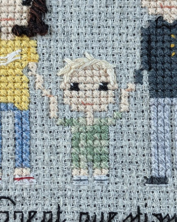 A cross-stitch and hand embroidered depiction of a little boy in a camo coloured outfit. He has messy, blonde hair and is holding his parents hands.
