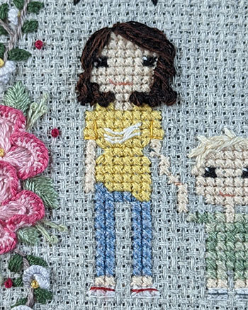 A cross-stitch and hand embroidered depiction of a woman with brown curly hair, a yellow tshirt and demin pants.