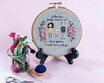 A setup display of the finished, hooped stitch people portrait. There are three people and beside them is hand embroidered flowers. Around the piece is floss and stitching supplies. The piece is upright in a stand.