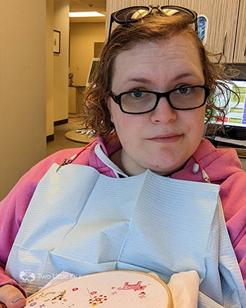 Selfie photo of Kate sitting in a dentist chair, looking a little exasperated. You can see at the bottom of the photo she's working on a cross-stitch. She's wearing the dentist's sunglasses on her head and a paper clip-on bib, and the dentist office can be seen behind her.