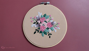 The entire hand embroidered floral piece. It is framed in an embroidery hoop and laying on a burgundy background. The floss colours are pinks, purples and greens.