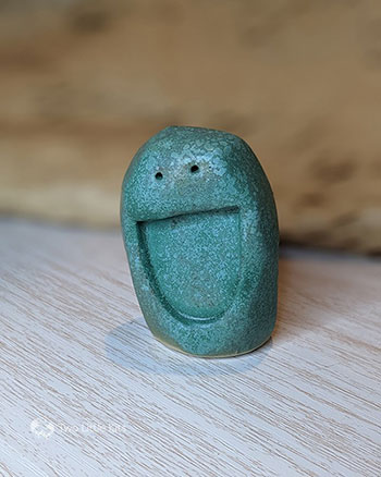 A green-glazed clay , tall 'blob' which is about the length of your palm, that's been stamped (before fired) with a semi-circle mouth and two tiny little circle eyes, so it looks like it has an enormous smiley face. The green glaze is mottled in appearance.