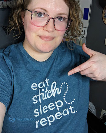 A selfie of Kate wearing one of her brand new t-shirt designs. She's wearing the teal colour variant. Kate is pointing at the t-shirt in a silly way and has a slight smirk on her face while looking at her phone.