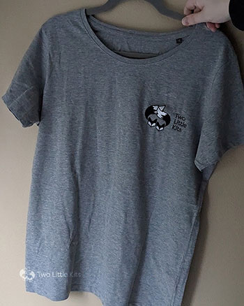 A photograph of a grey t-shirt on a hanger against a tan wall. On the left breast -area, there is a black and white machine embroidered graphic of the Two Little Kits logo.