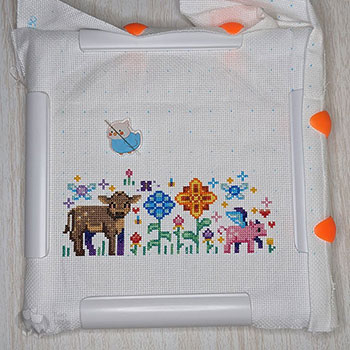 A colourful cross-stitch piece that depicts a cow, a pig with wings and lots of flowers.