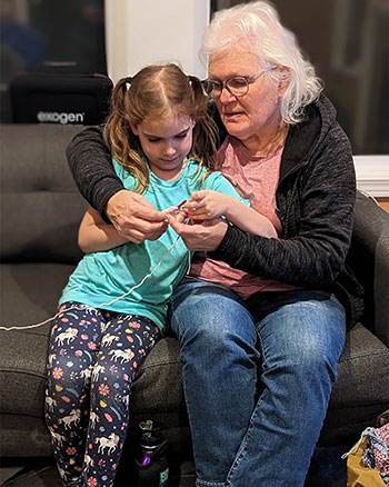 A grandmother and granddaughter sitting close together on a couch; the elder teaching the younger how to crochet.