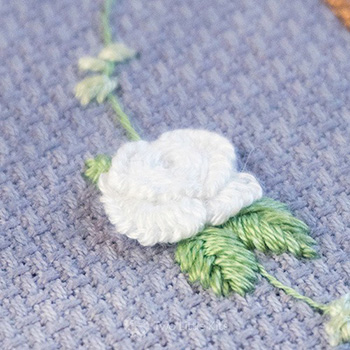 A close-up photo of a white flower on a green circular border with green flowers. It's all on a misty-blue fabric.