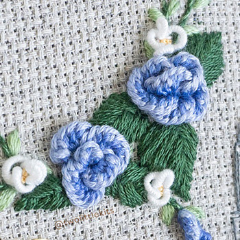 Two-toned blue cast-on stitched flowers and little bullion-stitched white flowers amongst green leafage, all hand stitched.